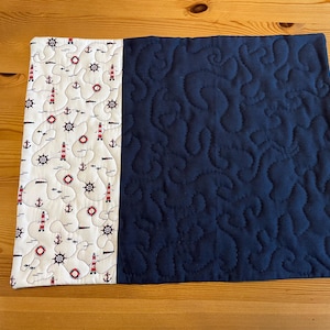 Könnte beinhalten: Ein rechteckiger, gesteppter Tischläufer. Eine Seite zeigt einen weißen Stoff mit roten Leuchttürmen, Ankern und Steuerrädern. Die andere Seite ist einfarbig marineblau mit einem Steppmuster. Maße: ca. 40 cm x 30 cm.