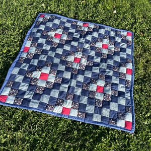Könnte beinhalten: Ein quadratischer Quilt mit einem Schachbrettmuster aus marineblauen, hellblauen und roten Quadraten. Einige Quadrate haben ein Blumenmuster. Der Quilt ist mit einem einfarbigen blauen Stoff eingefasst und liegt auf grünem Gras.