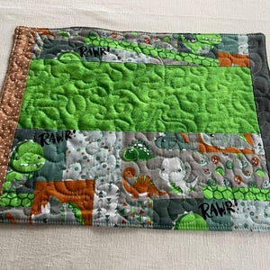 Könnte beinhalten: Ein rechteckiger, gesteppter Tischset mit Dinosaurier-Motiv. Es hat ein Patchwork-Design mit einer grünen Mitte und Dinosaurier-Prints. Das Wort "RAWR!" ist sichtbar. Die Ränder sind braun und grau.