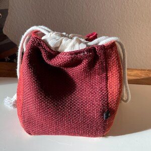 Könnte beinhalten: Ein kleiner, weinroter Kordelzugbeutel mit strukturiertem Gewebe. Die Tasche hat einen weißen Kordelzugverschluss und ein weißes Innenfutter. Die Tasche steht auf einer weißen Oberfläche.