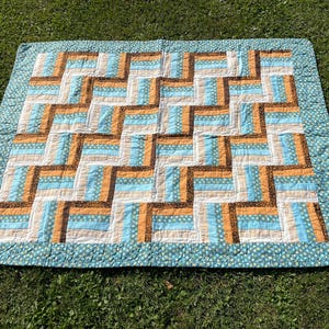 Könnte beinhalten: Eine Patchwork-Decke mit einem geometrischen Muster in Blau, Braun und Orange. Die Decke besteht aus Quadraten und Rechtecken, und die Farben sind in einem sich wiederholenden Muster angeordnet.