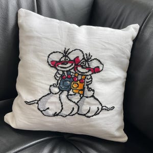 Könnte beinhalten: Weißes quadratisches Kissen mit einem Kreuzstichmuster von zwei Zeichentrickmäusen. Die Mäuse tragen Latzhosen und haben rote Akzente. Das Kissen liegt auf einer schwarzen Lederoberfläche.