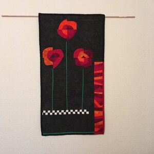 Könnte beinhalten: Ein schwarzer Wandbehang aus Stoff mit drei roten, orangefarbenen und gelben Blumen mit grünen Stielen. Die Blumen sind in einer Reihe angeordnet, mit einem schwarz-weißen Schachbrettmuster am unteren Rand und einem rot-orange-gelben Streifenmuster auf der rechten Seite.