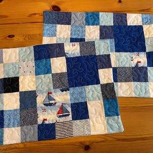 Könnte beinhalten: Zwei nautisch thematisierte, gesteppte Tischsets. Jedes Tischset hat ein Patchwork-Design in Blau- und Weißtönen. Ein Tischset hat Segelboot-Applikationen. Das andere hat eine Anker- und Segelboot-Applikation. Die Tischsets liegen auf einer Holzoberfläche.
