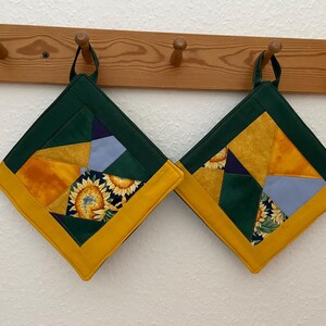 Könnte beinhalten: Zwei grüne und gelbe gequiltete Topflappen mit einem Sonnenblumenmuster, die an einem Holzgestell hängen.