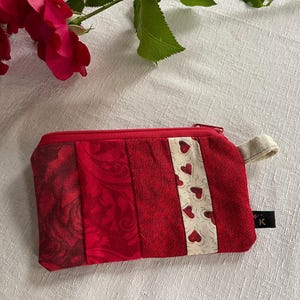 Könnte beinhalten: Ein rotes, gestepptes Münzfach mit Reißverschluss und einer kleinen Stoffschlaufe. Die Geldbörse hat ein Patchwork-Design mit verschiedenen roten Stoffen, darunter ein Abschnitt mit kleinen Herzmustern. Eine rote Rose ist im Hintergrund.