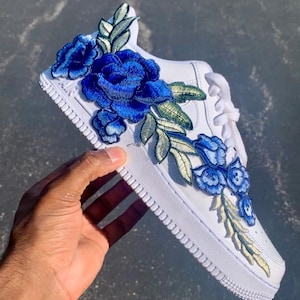 Pode incluir: Tênis brancos de couro com flores bordadas azuis. As flores estão em um padrão floral com folhas e caules.