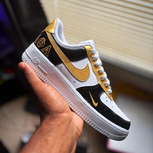 May include: White, black, and gold Air Force 1 sneakers. The shoe features a gold Nike swoosh, gold accents, and the letters ΦA on the side. The shoe has a white sole and white laces. The shoe is held in a person's hand.
