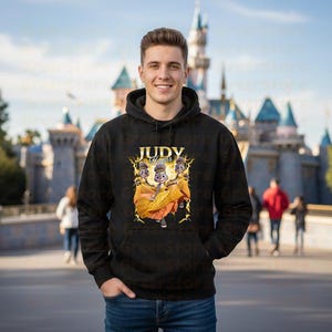 May include: Black hooded sweatshirt featuring a graphic of three cartoon characters with the text "Judy Hopps" in gold. The characters are wearing orange and yellow dresses. The background is a blurred image of a castle.