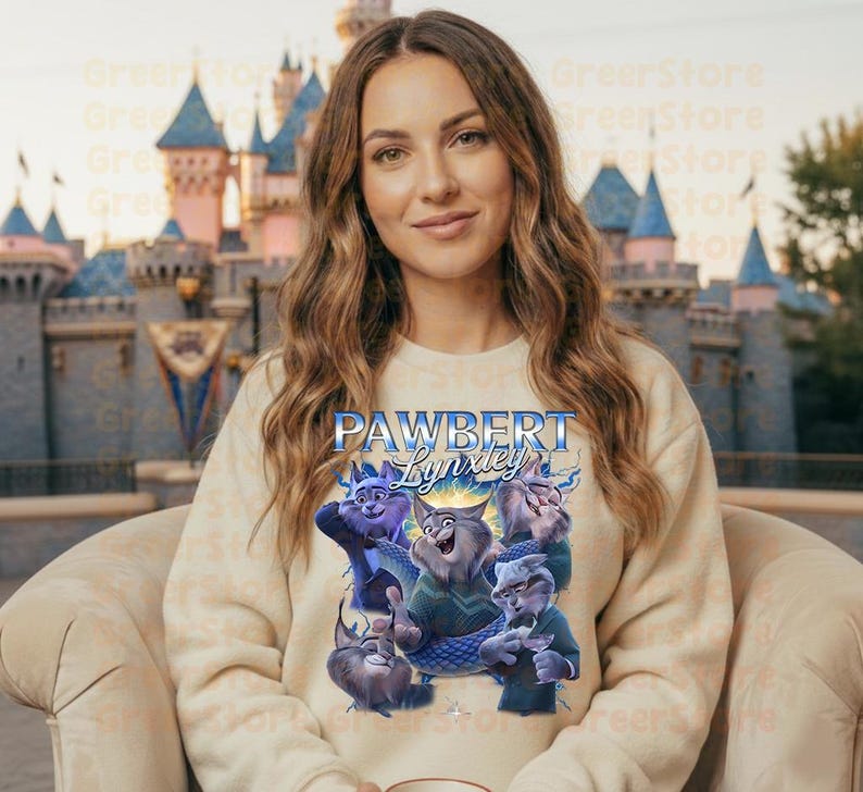 May include: A cream-coloured sweatshirt with a graphic of cartoon cats and the text "PAWBERT Lynxley". The cats are in various poses, with blue and purple accents. The background is a blurred image of a castle. The sweatshirt is a neutral colour.