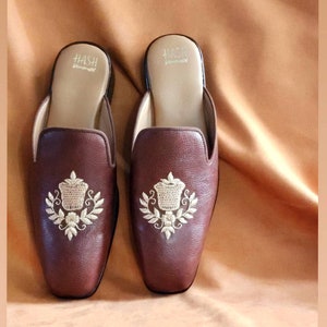 May include: A pair of brown leather slippers with gold embroidery featuring a floral design. The slippers have a pointed toe and a slip-on style. The brand name "HASH" is printed on the top of each slipper.