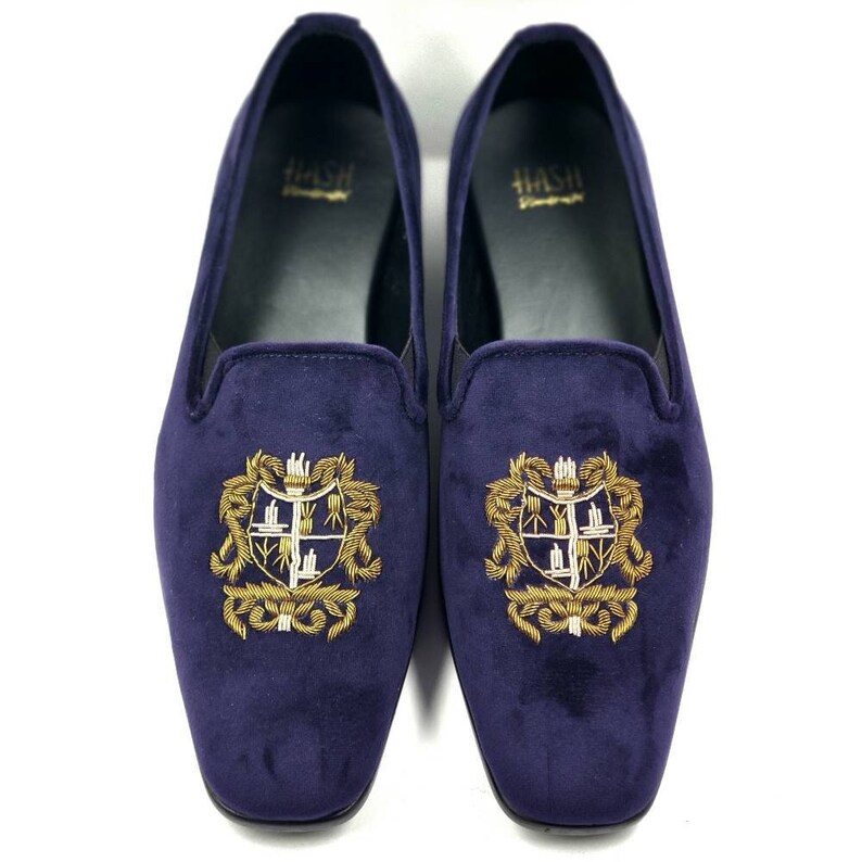 Puede incluir: Un par de zapatillas de terciopelo azul oscuro con crestas bordadas en oro. Las crestas presentan un escudo con una cruz y otros detalles.