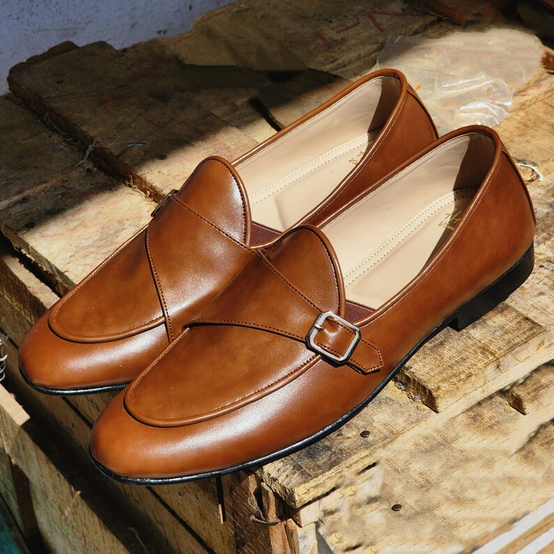May include: A pair of brown leather loafers with a single buckle strap. The loafers have a pointed toe and a low heel.