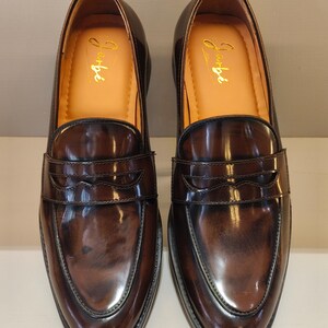 Pu&ograve; includere: Un paio di mocassini in pelle marrone con finitura lucida. Le scarpe hanno un design classico con una fascia sul collo del piede e un'iscrizione dorata all'interno. Le suole sono nere.