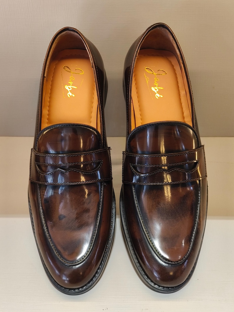 Pu&ograve; includere: Un paio di mocassini in pelle marrone con finitura lucida. Le scarpe hanno un design classico con una fascia sul collo del piede e un'iscrizione dorata all'interno. Le suole sono nere.