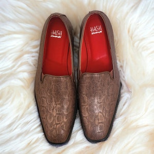 May include: A pair of brown leather loafers with a snakeskin pattern. The loafers have a red interior and the brand name "HASH" is printed on the inside of each shoe.
