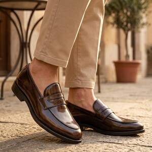 Pu&ograve; includere: Mocassini in pelle marrone lucida. Queste eleganti scarpe presentano un design classico con un cinturino. Sono abbinati a pantaloni beige, adatti a uno stile casual elegante o business.