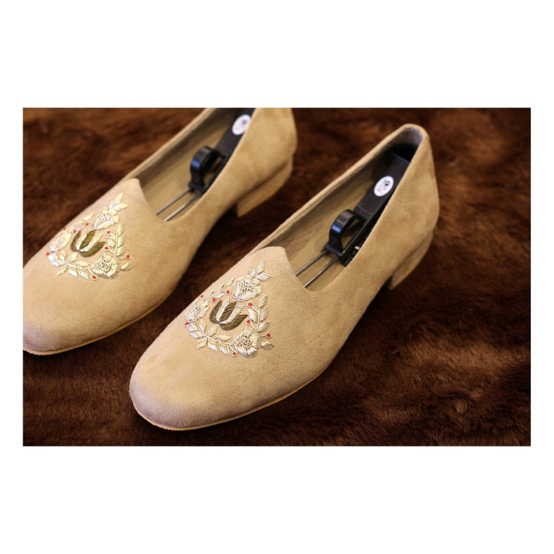 May include: A pair of beige suede slippers with gold and red embroidery. The embroidery features a floral design with a stylized letter "U" in the center.