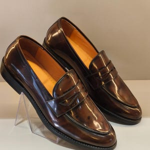 Pu&ograve; includere: Un paio di mocassini penny in pelle marrone con finitura lucida. Le scarpe hanno un design classico con una fascia sul collo del piede e una suola nera. L'interno &egrave; di colore marrone chiaro. Le scarpe sono esposte su un supporto trasparente.