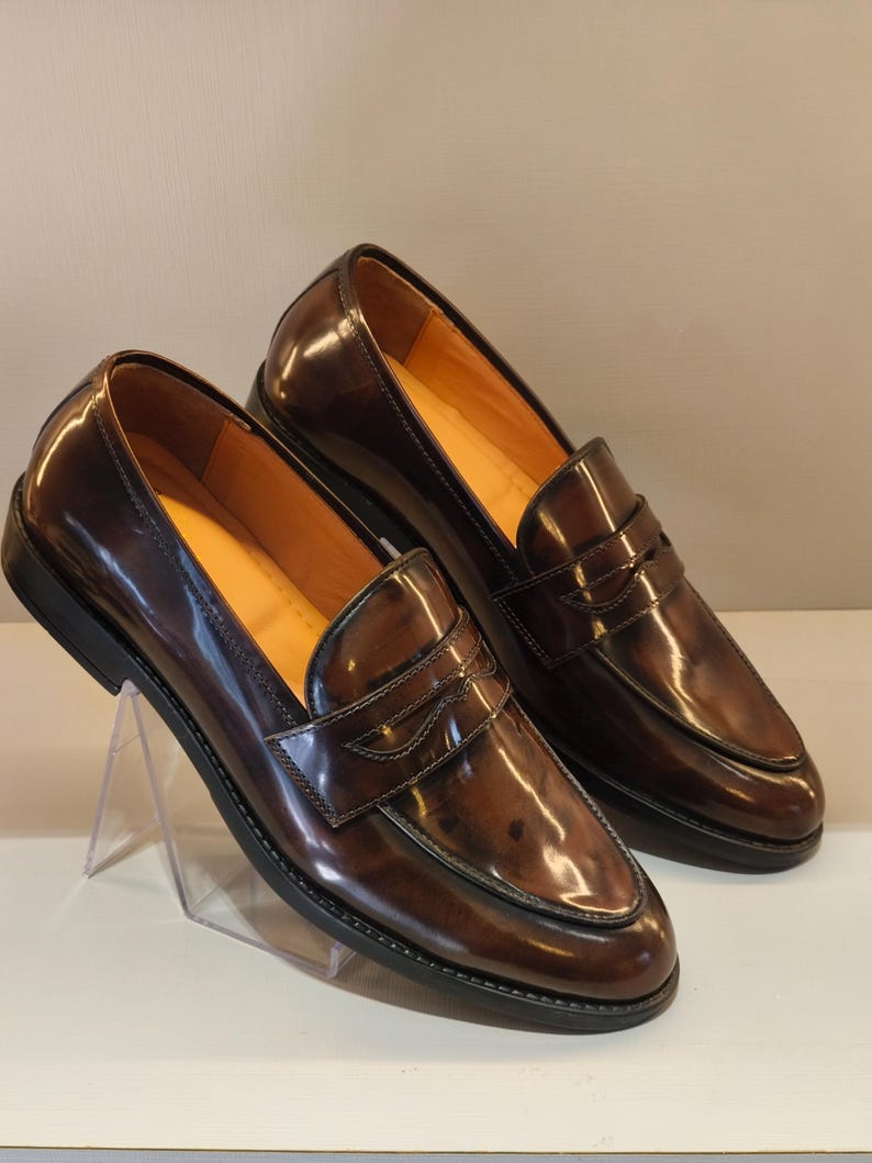 Pu&ograve; includere: Un paio di mocassini penny in pelle marrone con finitura lucida. Le scarpe hanno un design classico con una fascia sul collo del piede e una suola nera. L'interno &egrave; di colore marrone chiaro. Le scarpe sono esposte su un supporto trasparente.
