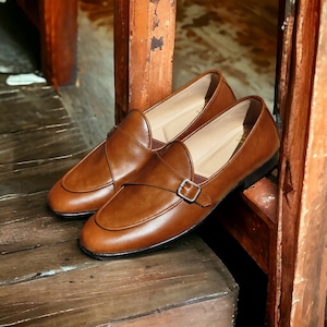 May include: A pair of brown leather monk strap shoes with a buckle closure. The shoes are sitting on a wooden surface.