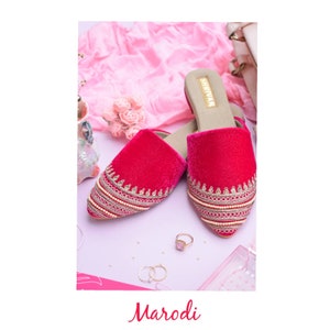 May include: A pair of pink velvet slippers with gold embroidery and pearl accents. The slippers have a pointed toe and a low heel. The brand name "SHIKVAA" is printed on the insole.
