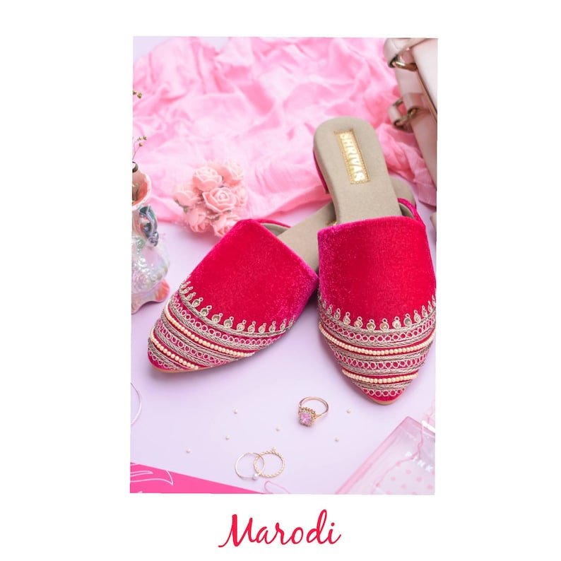 May include: A pair of pink velvet slippers with gold embroidery and pearl accents. The slippers have a pointed toe and a low heel. The brand name "SHIKVAA" is printed on the insole.