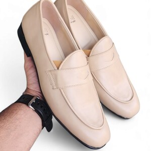 May include: A pair of beige leather loafers with a black sole. The loafers have a simple design with a single strap across the top.
