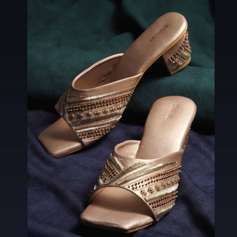 May include: A pair of rose gold heeled sandals with intricate embroidery and beaded embellishments. The sandals have a square toe and a block heel.