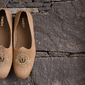 May include: A pair of tan suede loafers with gold embroidery. The loafers have the word "HOSH" written on the inside of each shoe.