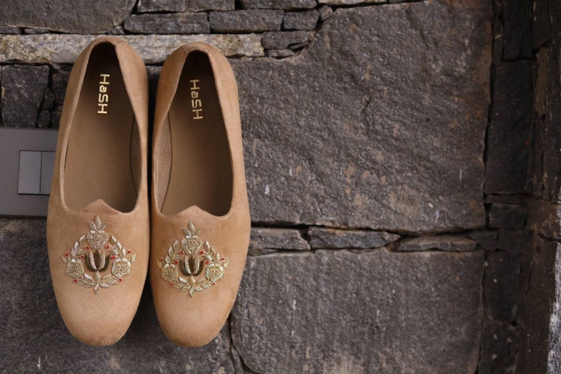 May include: A pair of tan suede loafers with gold embroidery. The loafers have the word "HOSH" written on the inside of each shoe.