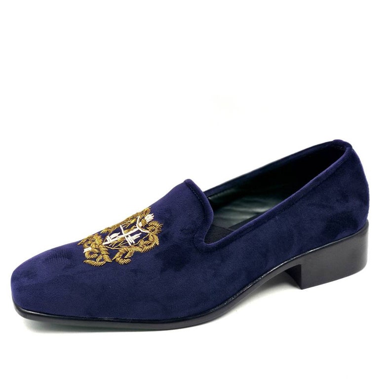 Puede incluir: Zapatillas de terciopelo azul marino con un escudo bordado en oro en la parte superior.