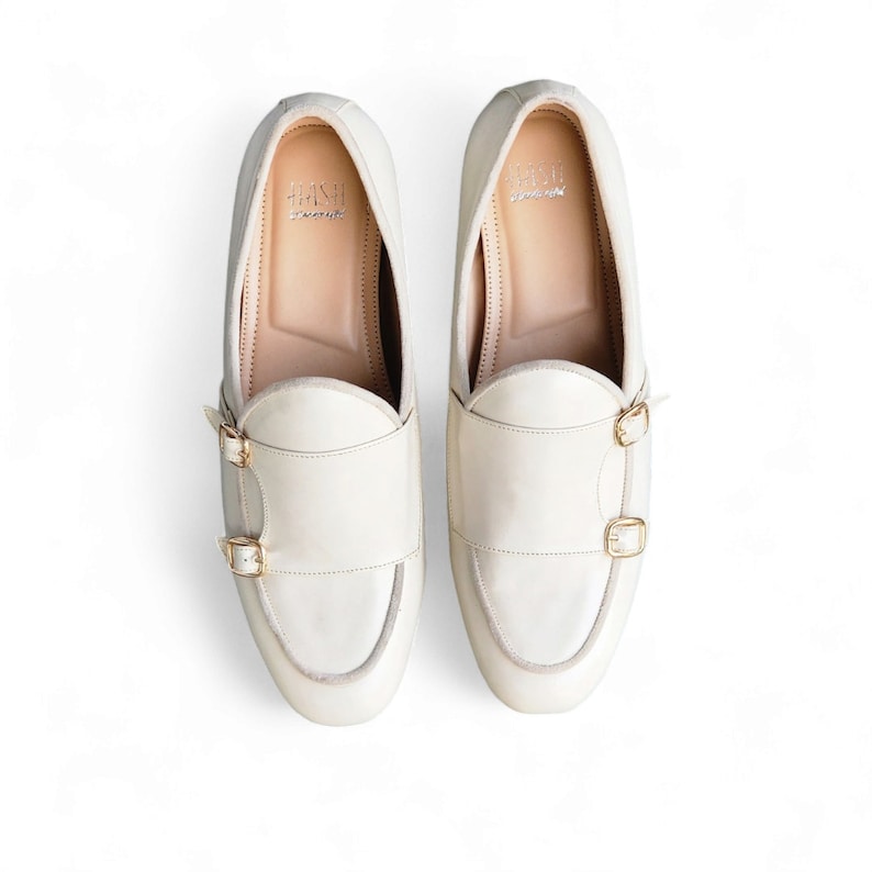 May include: A pair of off-white leather monk strap shoes with gold buckles. The shoes have a slightly pointed toe and a low heel.