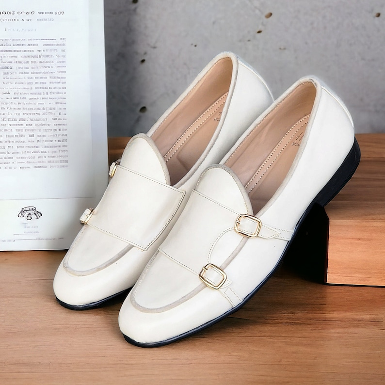 May include: A pair of off-white leather loafers with double buckles. The loafers have a rounded toe and a low heel.