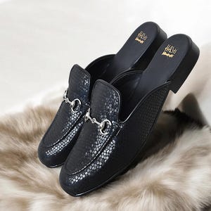 Puede incluir: Un par de zapatos negros de cuero con un patr&oacute;n de piel de serpiente y herrajes plateados. Los zapatos tienen el texto "HASH" impreso en la parte posterior.