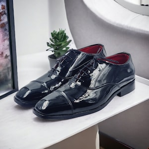 May include: A pair of black leather lace-up shoes with a cap toe design. The shoes have a glossy finish and red lining.
