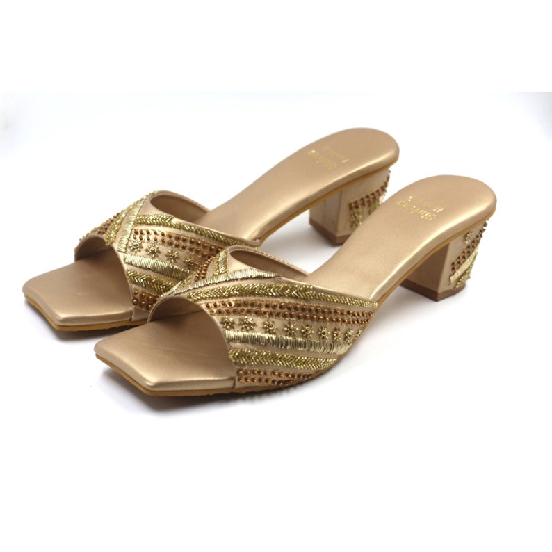 May include: A pair of gold heeled sandals with intricate embroidery and embellishments. The sandals have a square toe and a block heel.