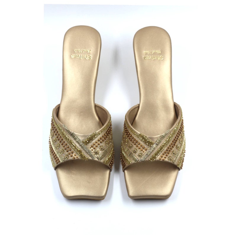 May include: A pair of gold heeled sandals with a square toe and intricate beaded embroidery. The sandals are labeled "SHRIYA CRAFTS".