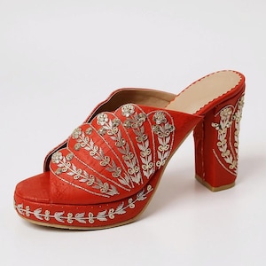 May include: Red platform heels with intricate silver embroidery. The open-toe mules feature a block heel and are adorned with floral and leaf patterns. The platform and heel are also embellished with silver embroidery.