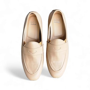 May include: A pair of beige leather loafers with a subtle heart-shaped design on the front. The loafers have a simple, classic style and are made by the brand "HASH"