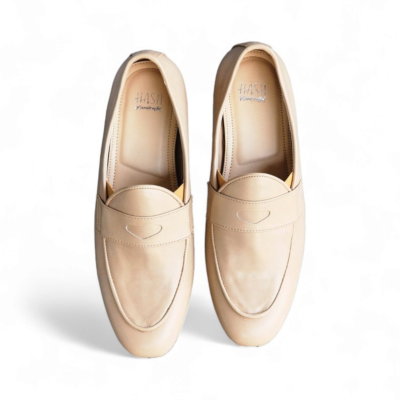 May include: A pair of beige leather loafers with a subtle heart-shaped design on the front. The loafers have a simple, classic style and are made by the brand "HASH"