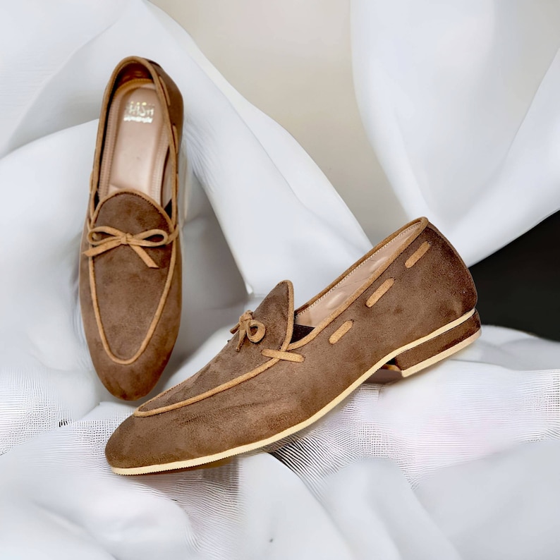 Pu&ograve; includere: Un paio di mocassini in camoscio marrone con un dettaglio a fiocco e un bordo in pelle color cuoio. I mocassini sono su uno sfondo di tessuto bianco.