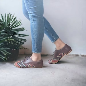 Puede incluir: Un par de zapatos de cuero marr&oacute;n con un patr&oacute;n tejido colorido en la punta y el tal&oacute;n. Los zapatos son usados por una persona que lleva jeans azules.