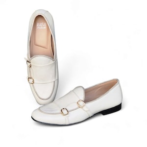 May include: A pair of white leather loafers with gold buckles. The loafers have a pointed toe and a low heel. The brand name "HASH" is visible on the inside of one of the shoes.