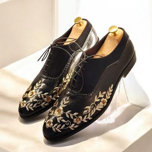 Può includere: Due scarpe in pelle nera con ricami floreali dorati. Le scarpe hanno lacci e sono su una superficie bianca.