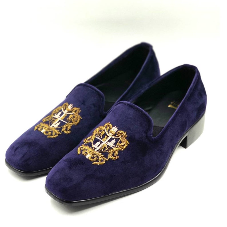 Puede incluir: Un par de mocasines de terciopelo azul oscuro con un escudo bordado en oro en cada zapato.