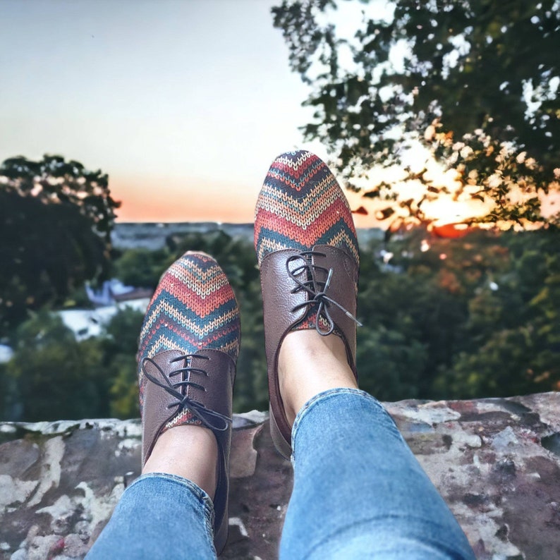 Puede incluir: Un par de zapatos de cuero marr&oacute;n con un patr&oacute;n geom&eacute;trico colorido en la parte superior. Los zapatos son usados por una persona que lleva jeans azules. Los zapatos est&aacute;n sentados en una superficie de piedra con un fondo borroso de &aacute;rboles y una puesta de sol.