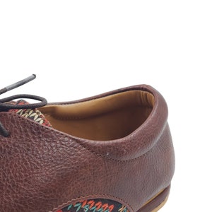 Puede incluir: Primer plano de un zapato de cuero marr&oacute;n con un detalle de tela estampada de colores en el tal&oacute;n y la punta. El zapato tiene un cierre de cordones y una suela de cuero marr&oacute;n.