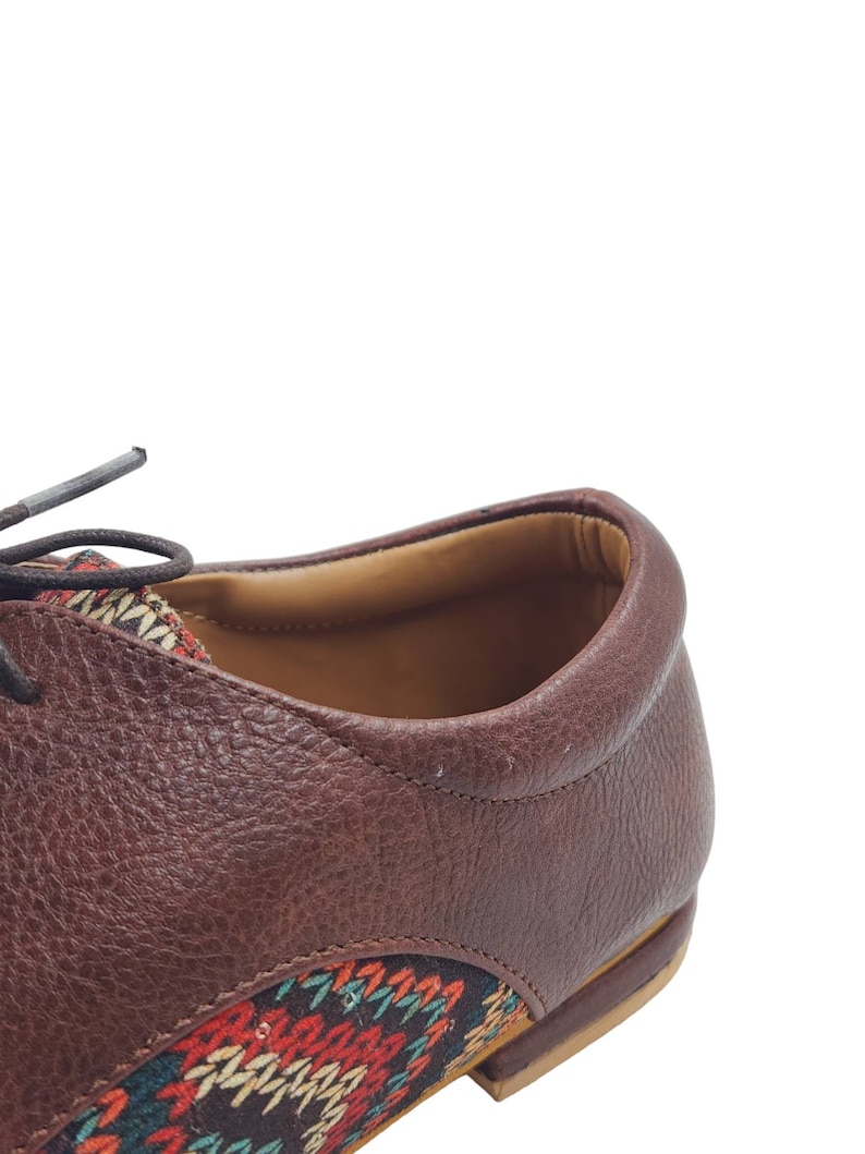 Puede incluir: Primer plano de un zapato de cuero marr&oacute;n con un detalle de tela estampada de colores en el tal&oacute;n y la punta. El zapato tiene un cierre de cordones y una suela de cuero marr&oacute;n.