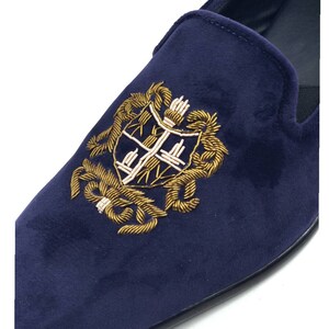 Puede incluir: Pantuflas de terciopelo azul marino con un escudo bordado en oro que presenta un escudo y detalles florales.